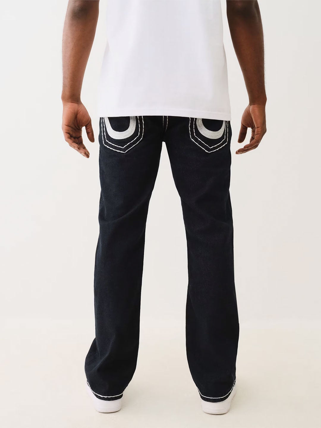 Back view of a model wearing black True Religion jeans with signature white contrast stitching on the pockets, paired with a white t-shirt and white sneakers, highlighting the brand's iconic denim design.