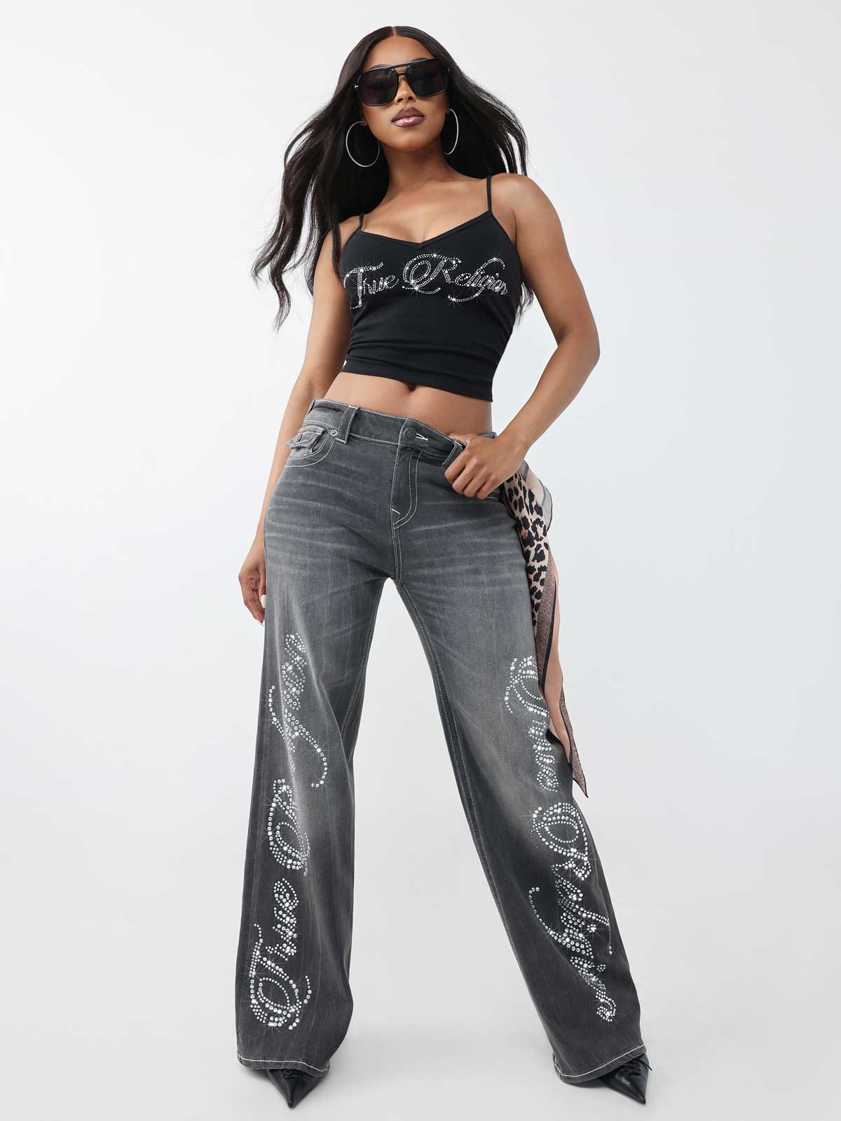 Woman wears black true religion sunglasses, black true religion tank top with crystal logo, and gray acid wash true religion jeans with crystal details