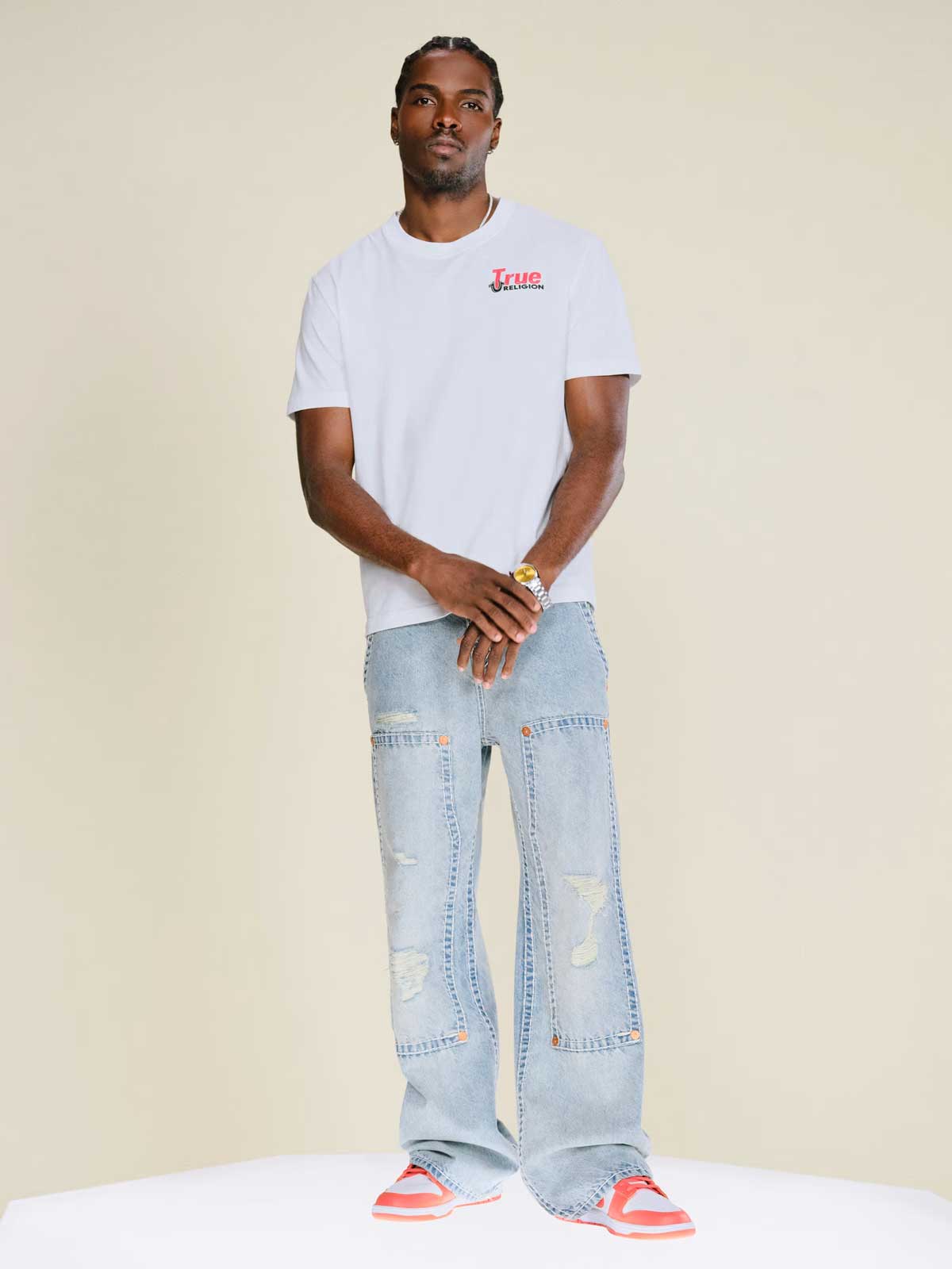 Man wears true religion light wash baggy jeans, red and white sneakers, and a true religion graphic tee