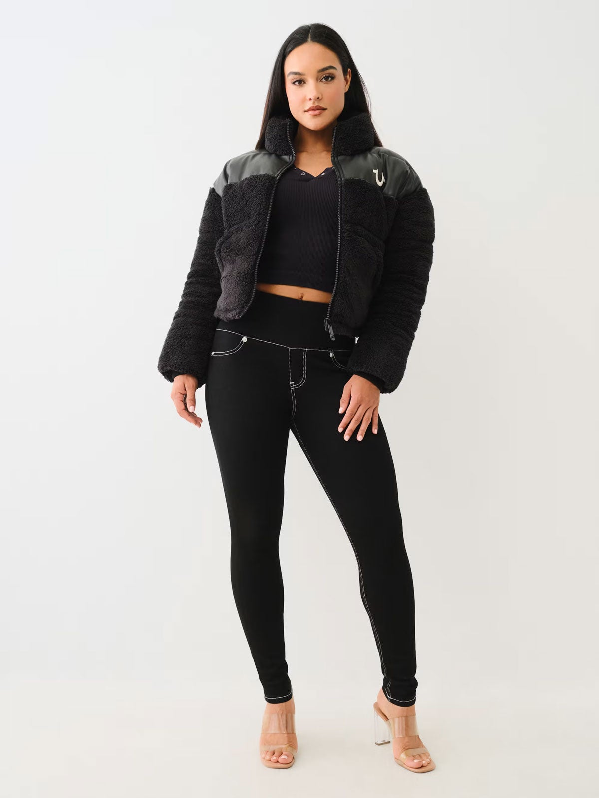 A woman stands against a light gray background wearing a cropped black fuzzy jacket, black leggings with contrast stitching, a black crop top, and clear high-heeled sandals. 