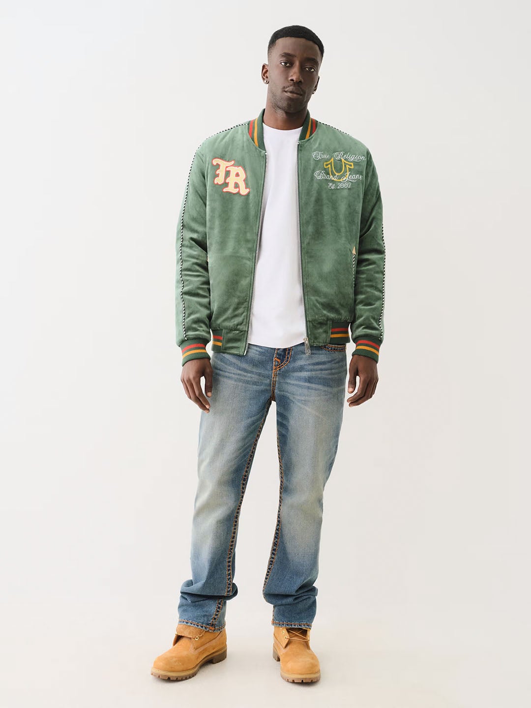 Model wearing a green True Religion varsity-style jacket with embroidered details, paired with a white t-shirt, light wash jeans, and tan work boots, showcasing a casual yet polished streetwear look.