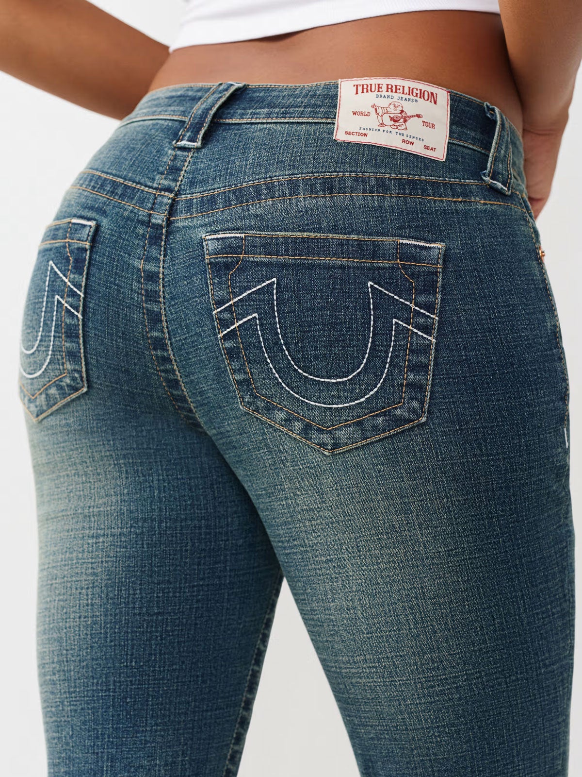 Close-up back view of a person wearing blue True Religion jeans, showing the brand patch at the waistband and white-stitched flap back pockets.