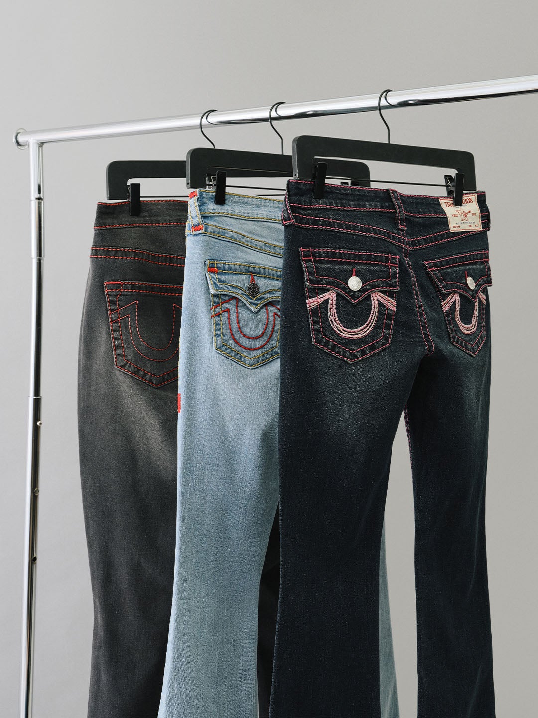 Three pairs of true religion jeans on hangers with red horseshoe stitching on the back pockets.