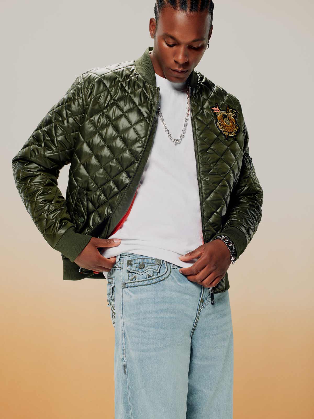 Man wears a true religion quilted green bomber jacket, a white tee, and true religion light wash jeans 