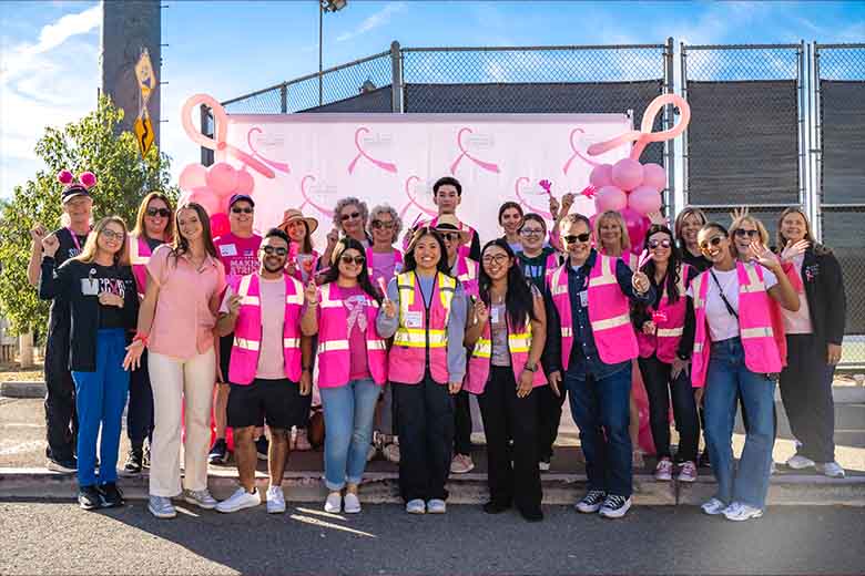 A group of True Religion volunteers and Breast Cancer Awareness volunteers wearing bright pink safety vests and casual clothing pose together in front of a banner featuring pink ribbons at a breast cancer awareness event. The cheerful group stands outdoors on a sunny day, surrounded by balloons and supportive decorations.