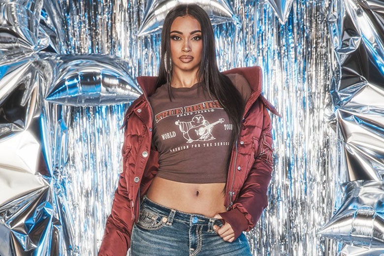Woman wearing a true religion red puffer jacket, a brown baby tee, and true religion jeans standing in front of a background of silver streamers and balloons