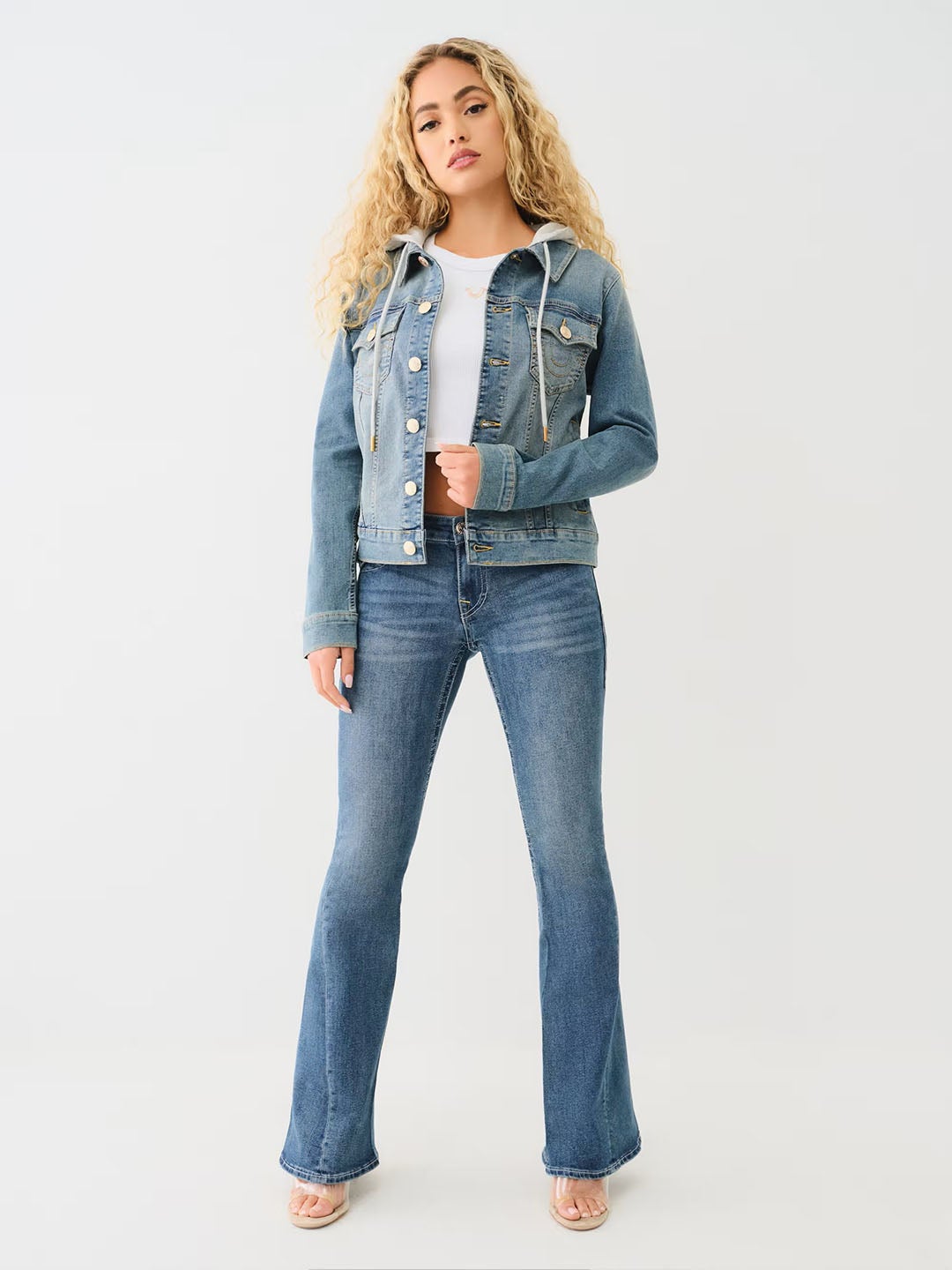 Woman wearing a light wash jean jacket with a hood, a white t shirt, and blue true religion jeans.