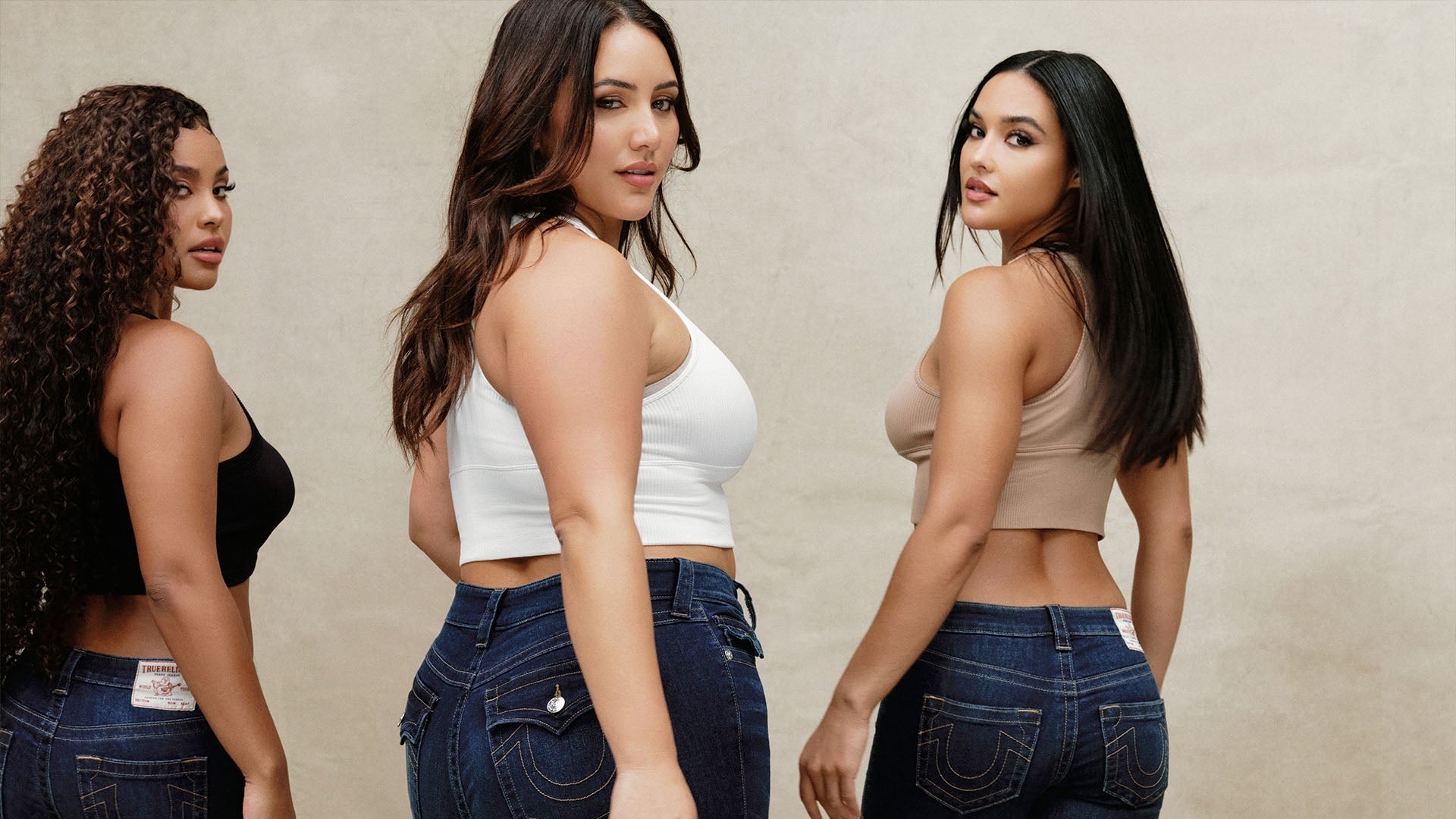 Three women in tank tops wearing different sizes of dark wash true religion jeans.