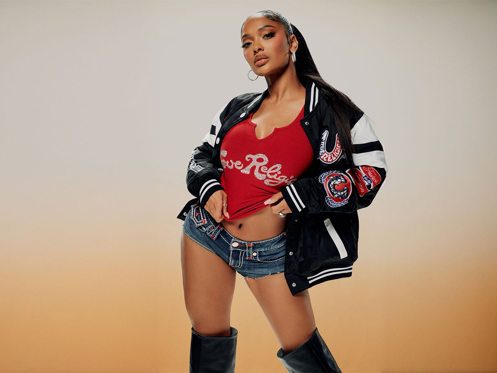 Woman wears true religion low rise jean shorts, a true religion red tee shirt, and a true religion black, white, and red varsity jacket 