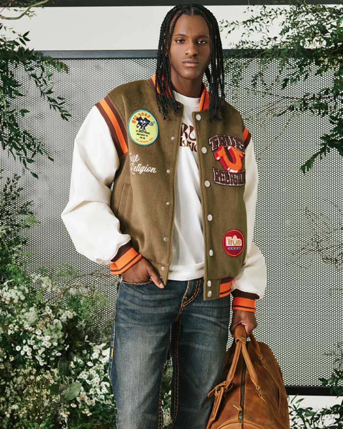 A man wearing a brown-and-cream varsity jacket, white shirt, and blue jeans stands in front of a lush green floral backdrop, holding a brown duffel bag.