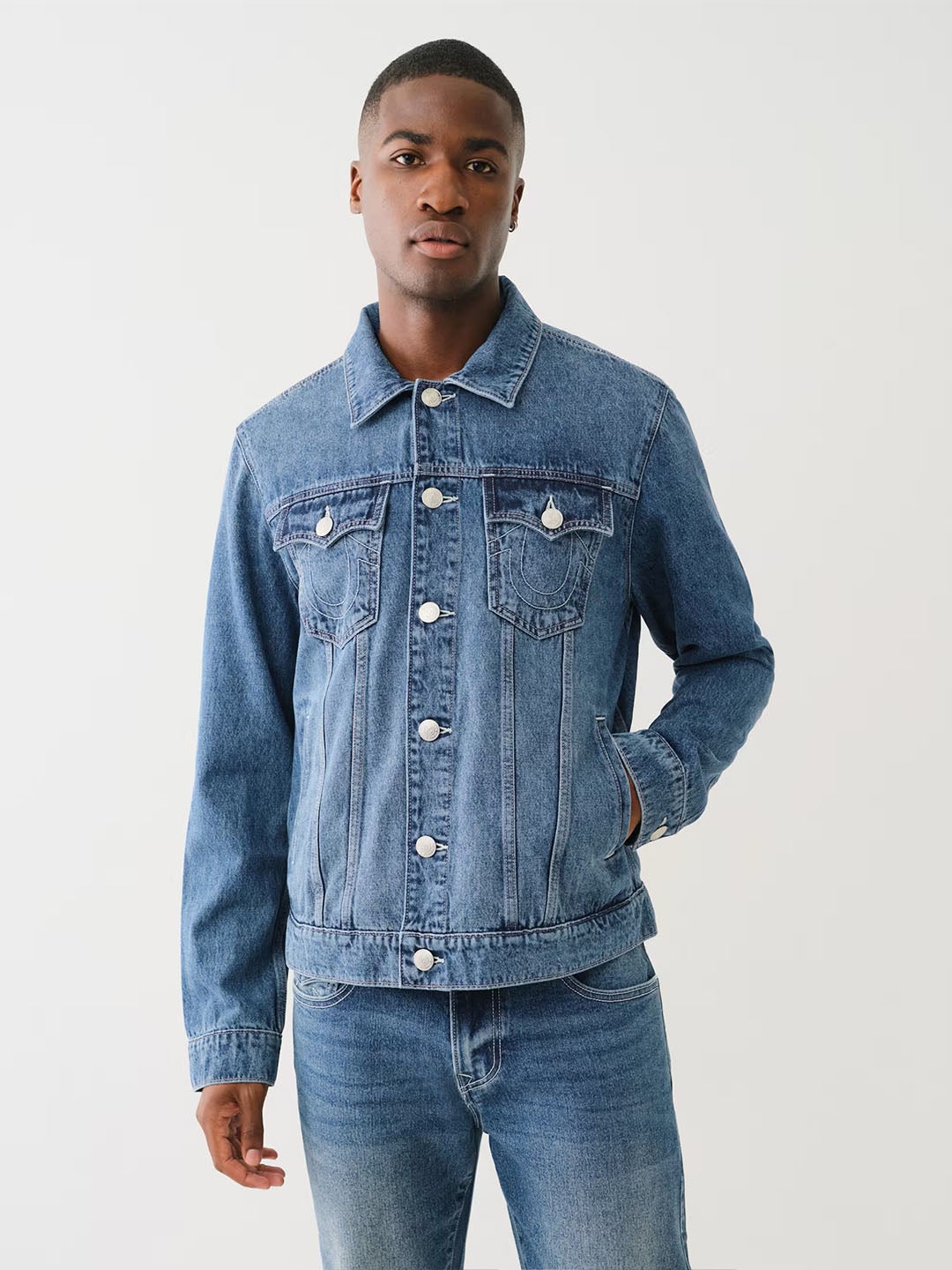 Man wears a blue true religion denim jacket with silver buttons and blue jeans.