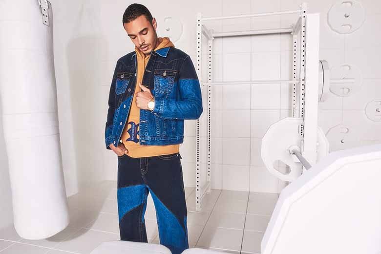 Man poses against a white industrial background wearing a denim jacket, brown sweatshirt, and light and dark wash pattern jeans.