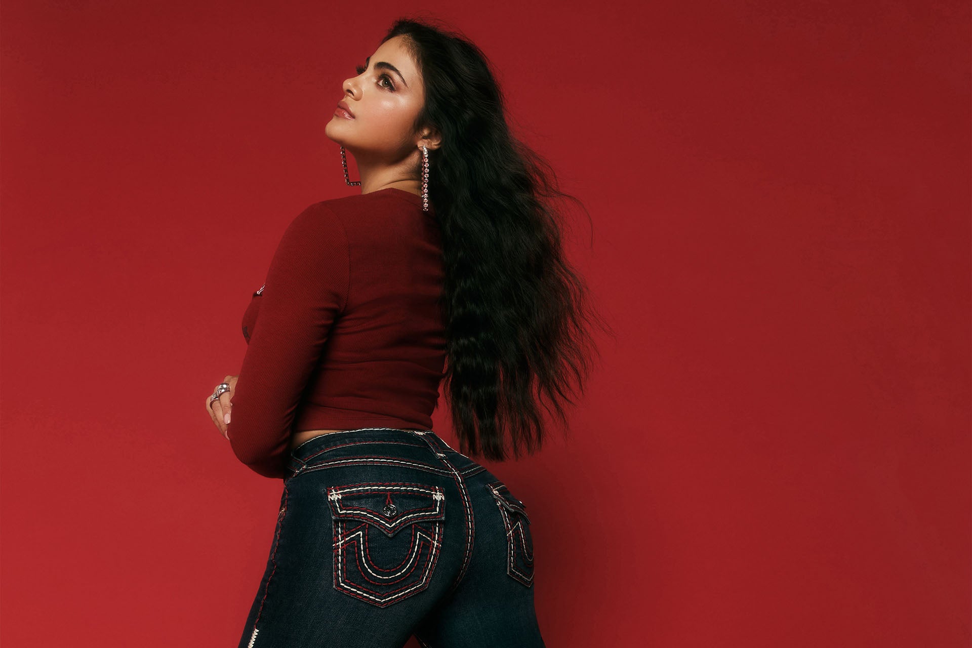 Woman that is kneeling, wearing dark wash true religion jeans with white and black stitching against a red background.