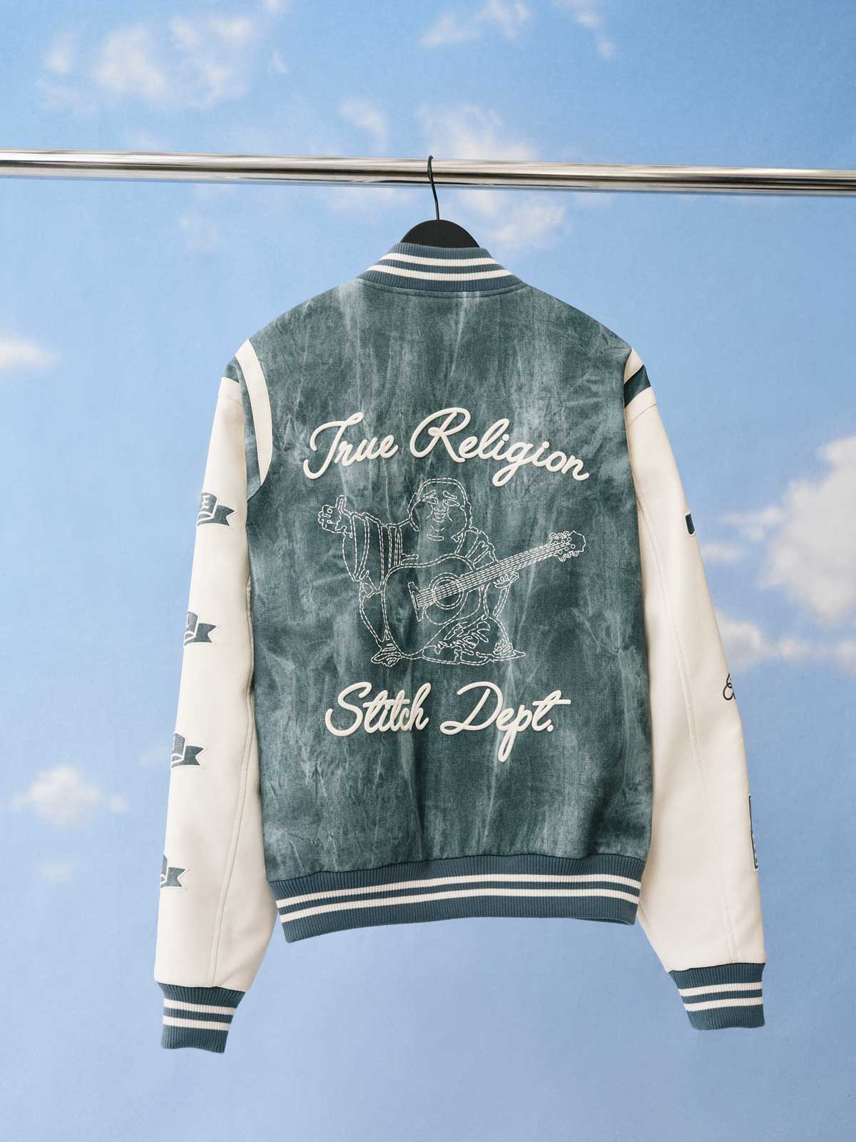 The back of a denim blue and white varsity jacket hangs on a clothing rack against a sky-blue background.