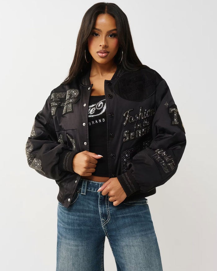 A woman with long dark hair poses against a light gray background wearing hoop earrings, a black embellished bomber jacket, a black top, and blue jeans.