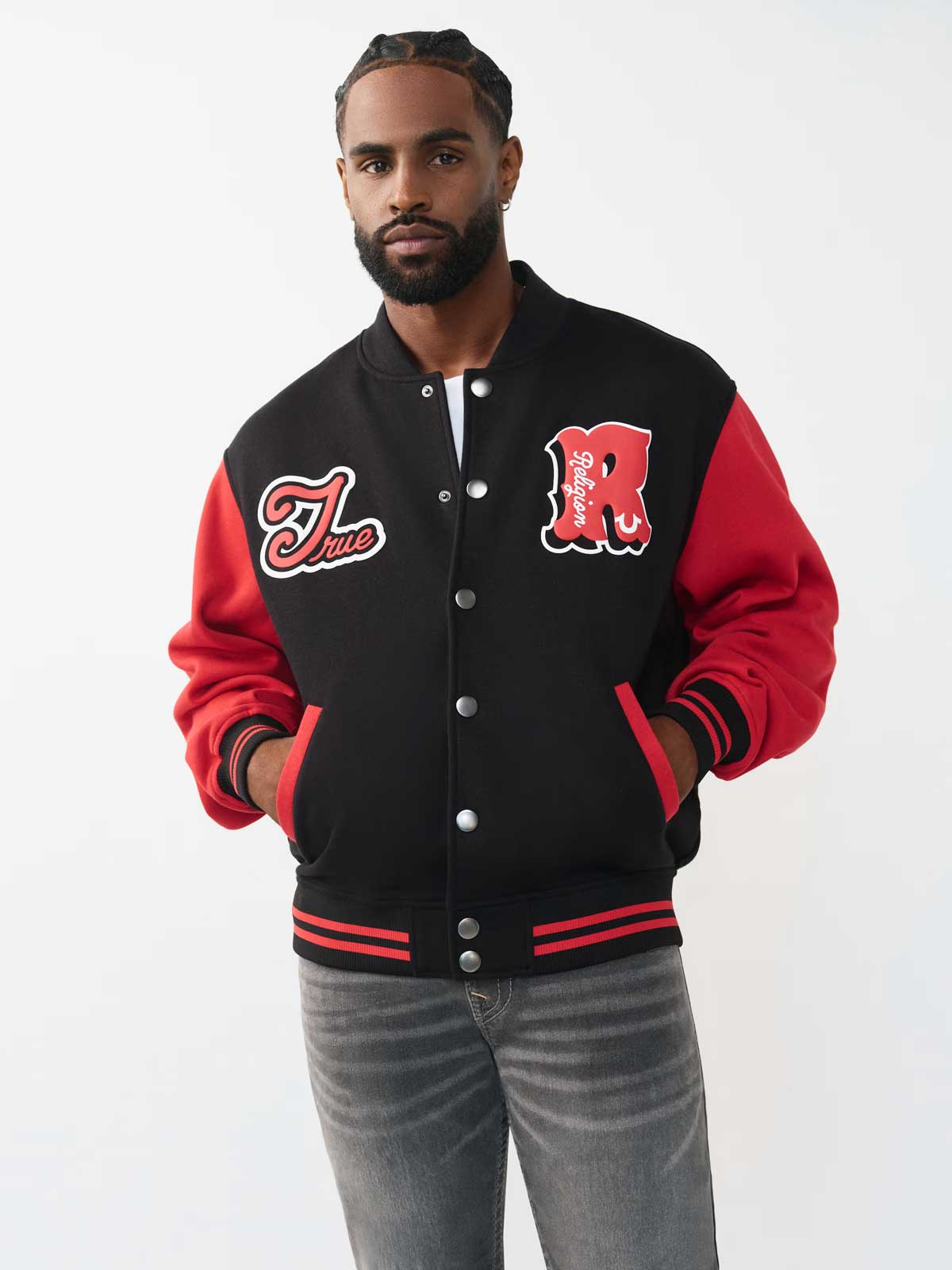 A man with braided hair and a beard poses against a light gray background wearing a black varsity jacket with red sleeves, gray jeans, and his hands in the jacket pockets.