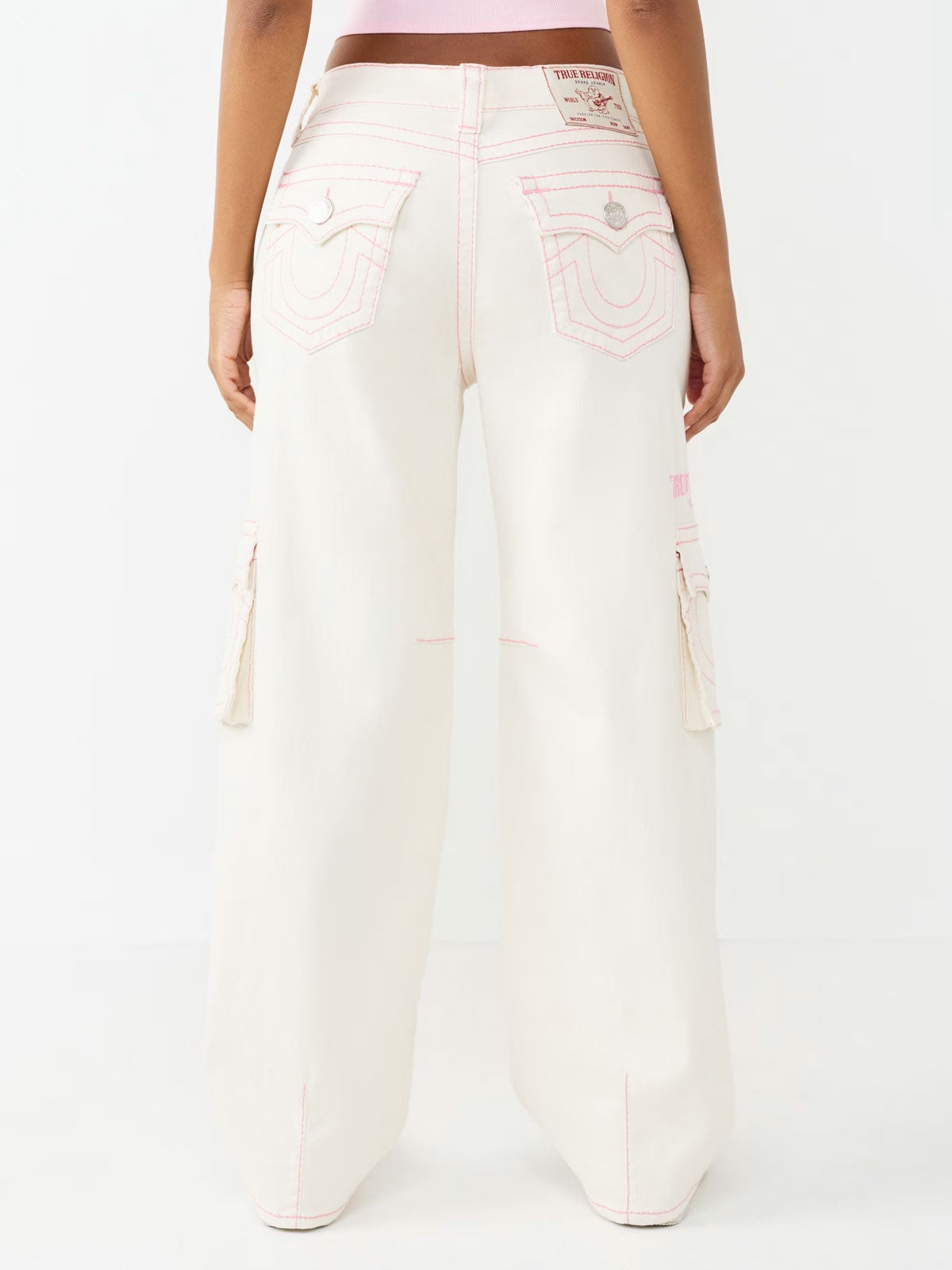back view of a woman’s legs standing in front of a white backdrop wearing white true religion jeans with pink stitching