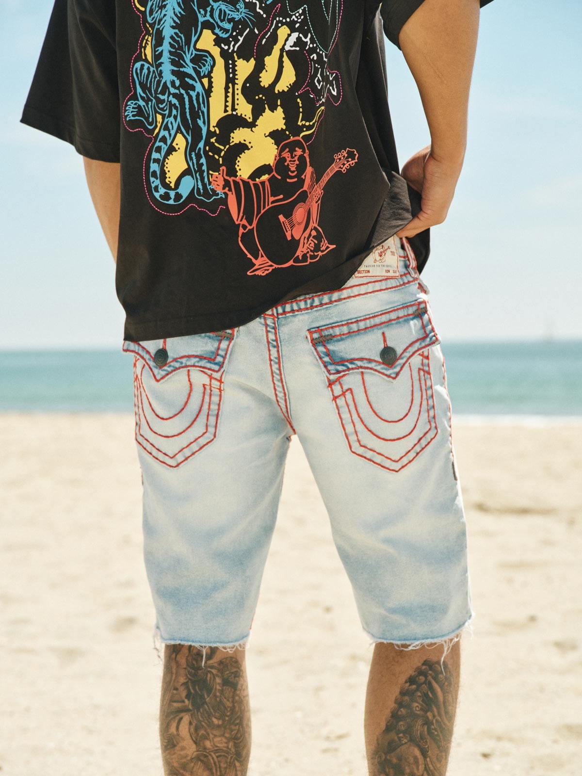 Back view of a man’s legs standing on a beach wearing baggy denim shorts and a graphic black t-shirt